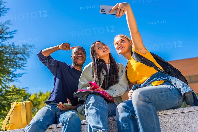 Group of smiling multi-ethnic students taking selfie sitting outside the campus in a sunny day