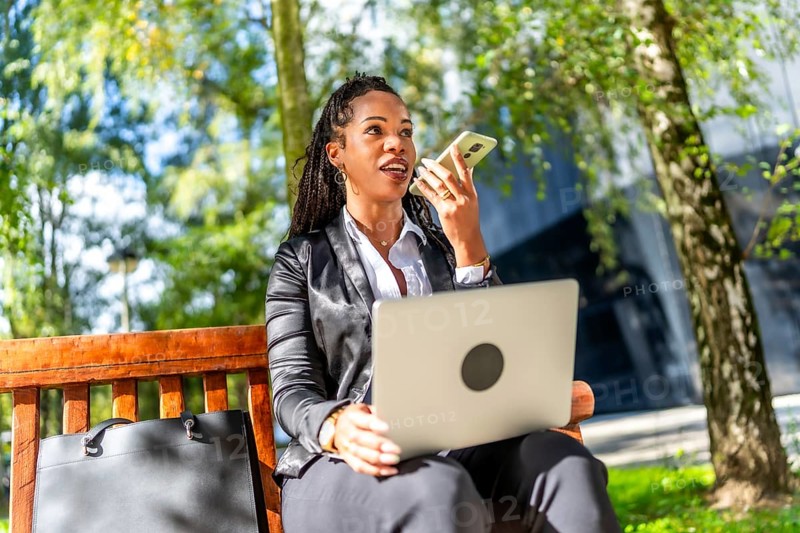 Latin young businesswoman sending voice message with mobile phone while working with laptop sitting on a bench outdoors