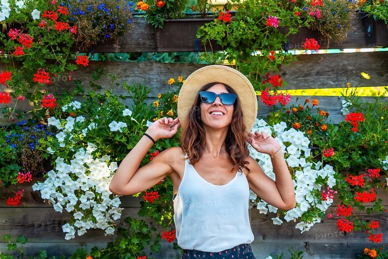 A woman wearing a straw hat and sunglasses is standing in front of a wall of flowers. She is smiling and she is enjoying the beautiful display