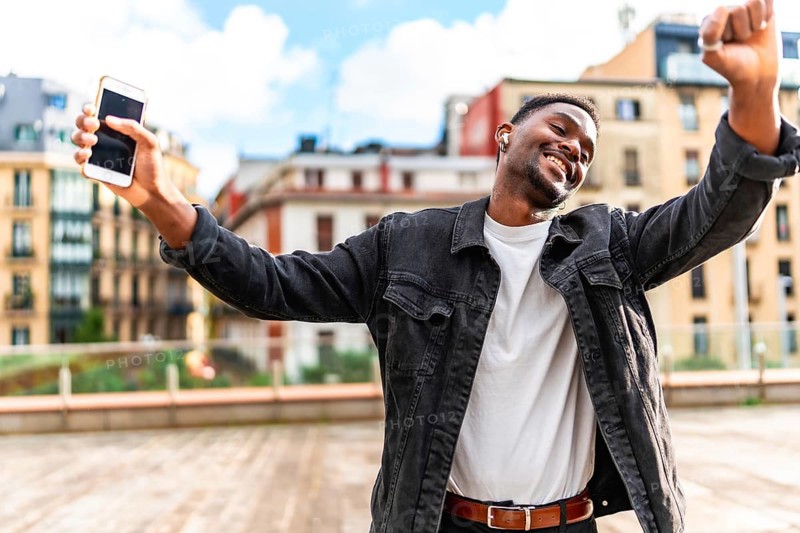 African man dancing happy while listening to music using mobile phone and wireless earbuds in the city