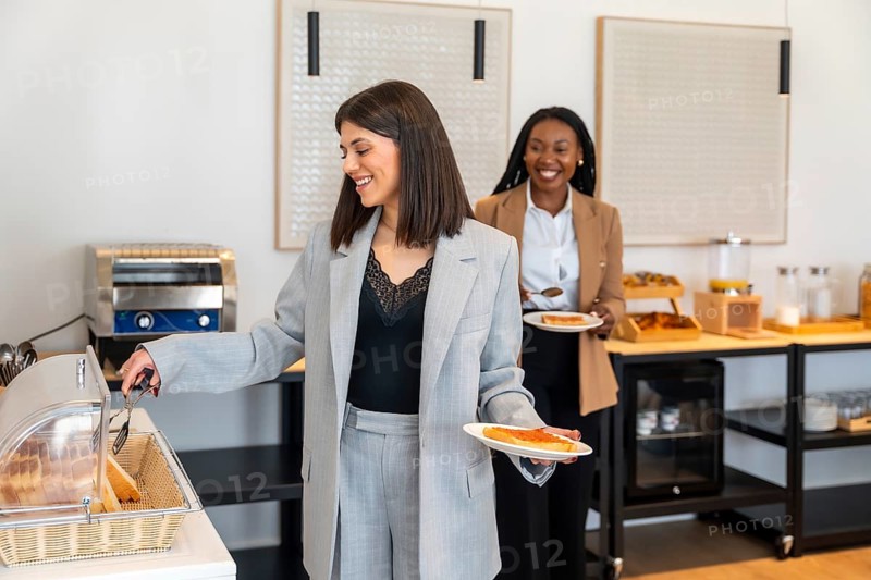 Two businesswomen are serving themselves at a hotel breakfast buffet, enjoying toast and other morning treats during their business trip