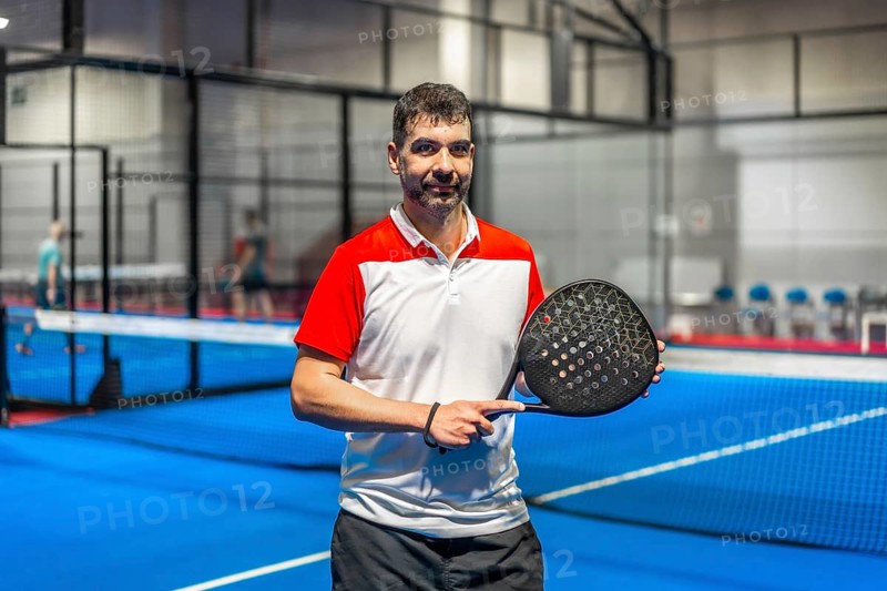 Portrait of a sportive mature man with paddle tennis racket standing in a blue indoor paddle court