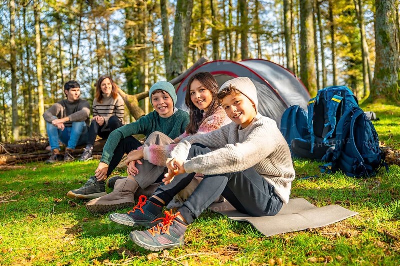 Three kids sitting next to their parents in the forest enjoying together a camping weekend