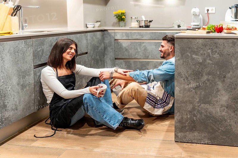 Two chefs are sitting on the floor of their modern kitchen, enjoying a coffee break and chatting after a busy cooking session