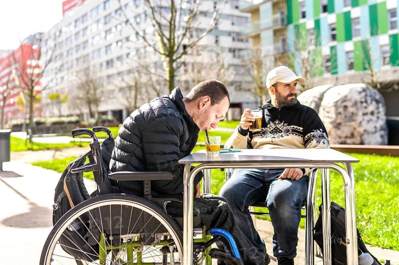 Two men enjoy drinks at an outdoor cafe, one using a wheelchair, demonstrating inclusivity and friendship