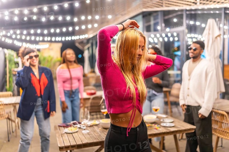 Blonde sensual woman dancing with friends at night party at the terrace of a bar decorated with lights