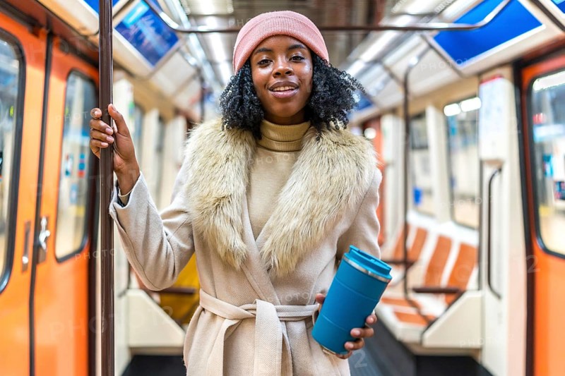 Smiling young black woman wearing winter coat and pink wool hat holding reusable coffee cup while riding subway train