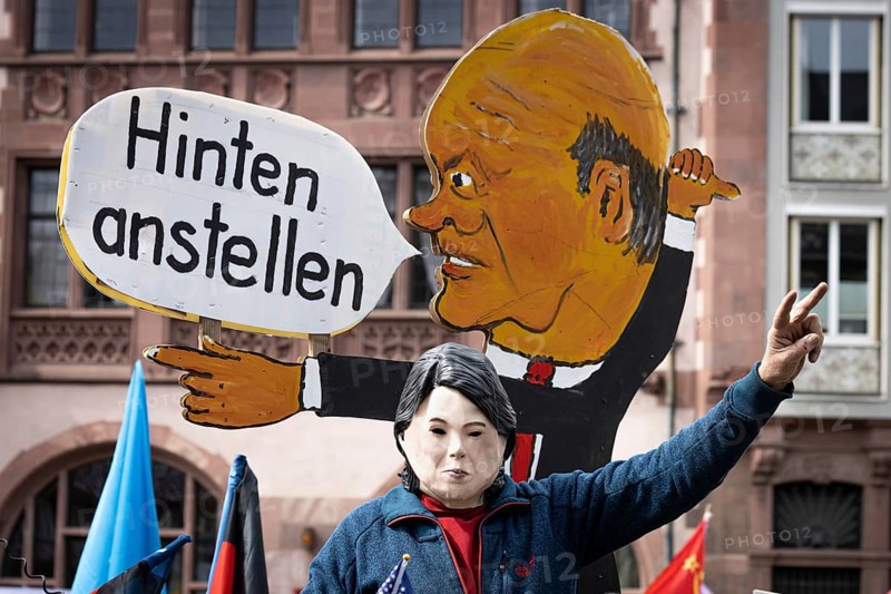 An activist wearing a mask of German Foreign Minister Annalena Baerbock sits on a replica of a tank in front of a speech bubble of German Chancellor Olaf Scholz during the rally for the Easter March on the Römerberg in Frankfurt am Main, Germany, on which it is written Anstellen in the back, meaning climate goals behind rearmament and energy security. A major theme of the rally was the war in Ukraine., Römerberg, Frankfurt am Main, Hesse, Germany, Europe