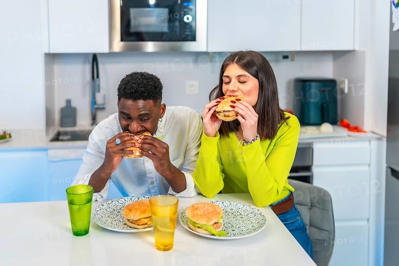 Young multiethnic couple eating homemade burgers in their modern kitchen, enjoying a casual meal together