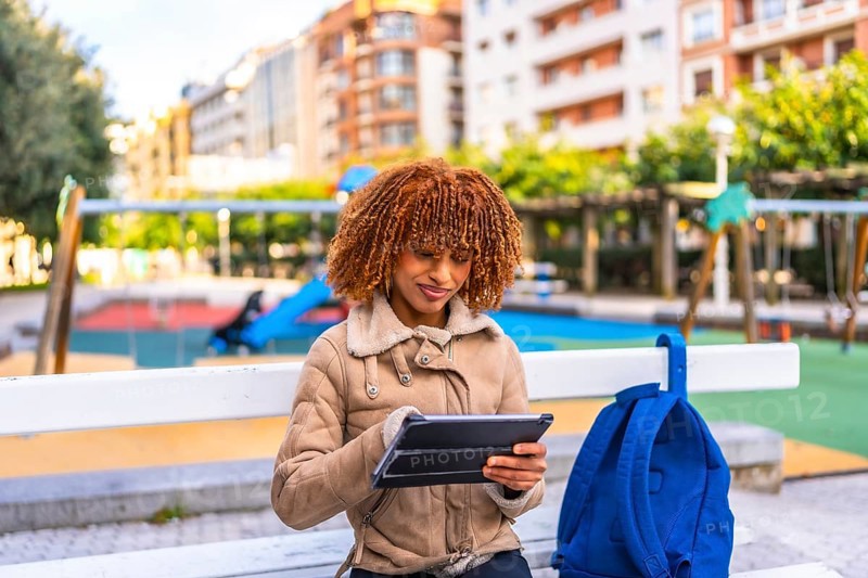 Latin young woman sitting on urban park bench using digital tablet