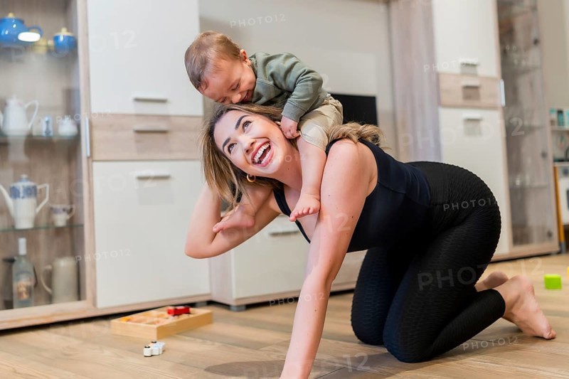 Young mother joyfully playing with her baby son, carrying him on her back while enjoying quality time together in the living room