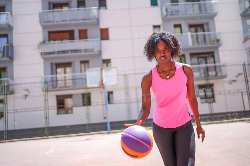Confident afro woman looking at camera while dribbling in an urban basketball court