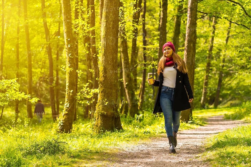 Latin girl with a black jacket, scarf and red woolen hat enjoying in an autumn park, walking along a path with a coffee at sunset