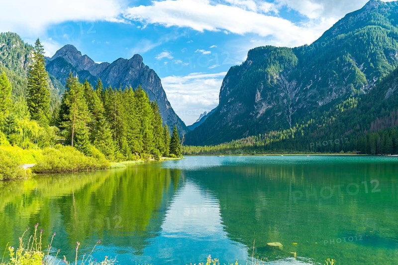 Stunning alpine landscape reflecting on lake dobbiaco's emerald waters, surrounded by lush vegetation and majestic peaks