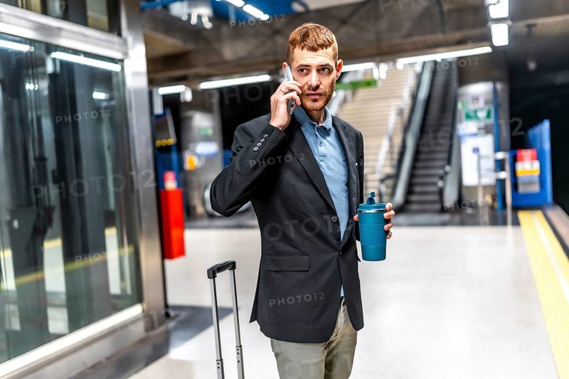 Businessman waiting for subway, talking on phone and holding reusable coffee cup, in a subway station