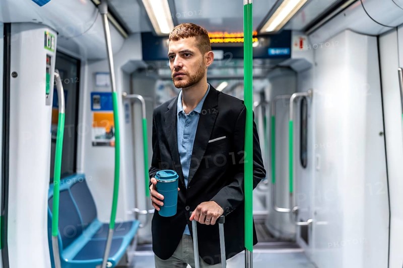 Young businessman commuting to work by subway, holding a reusable coffee cup and a trolley bag while looking out the window