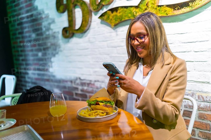 Beautiful latin woman wearing business casual clothes enjoying tasty burger and texting on the phone
