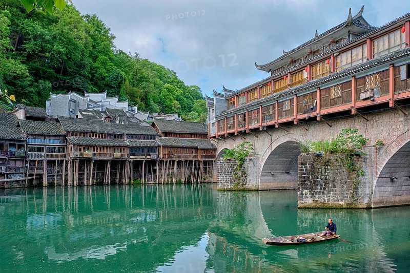FENGHUANG, CHINA, APRIL 23, 2018: Unidentified chinese man in boat Feng Huang Ancient Town (Phoenix Ancient Town) on Tuo Jiang River with bridge and tourist boat. Hunan Province, China, Asia