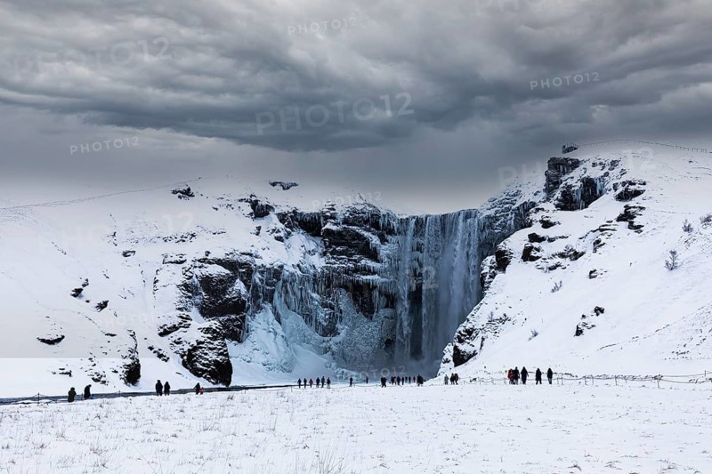 Tourists at Skogafoss waterfall, snowy and icy rock face, dark clouds above, Sudurland, Iceland, Europe