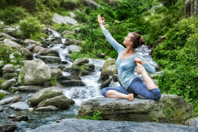 Young sporty fit woman doing yoga asana Eka pada rajakapotasana, one-legged king pigeon pose at tropical waterfall