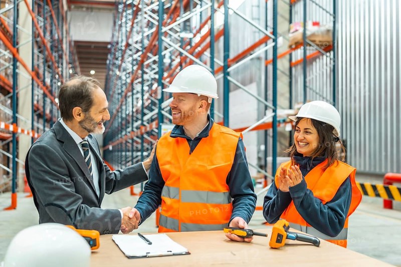 Manager shaking hands with a warehouse worker after closing a deal, his colleagues clapping hands, logistics warehouse interior