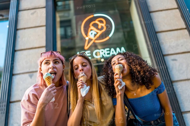 Low angle view photo of three diverse female friends enjoying eating a delicious ice cream outside the store