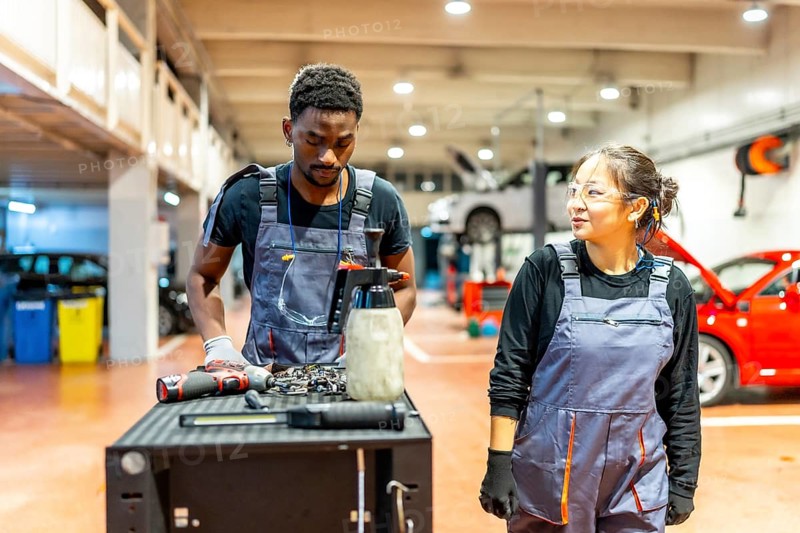 Two auto mechanics, a man and a woman, stand beside a workbench, reviewing tools and discussing car maintenance in a bustling garage