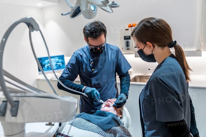 Dentist wearing magnifying glasses and face mask performing a dental procedure on a senior female patient assisted by a dental nurse