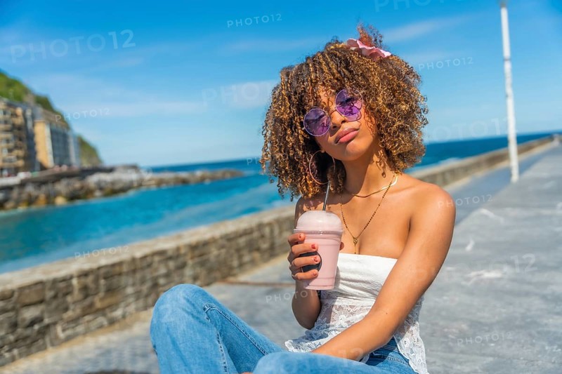 Cool latin young woman with curly hair in summer clothes drinking a shake outdoors next to the sea