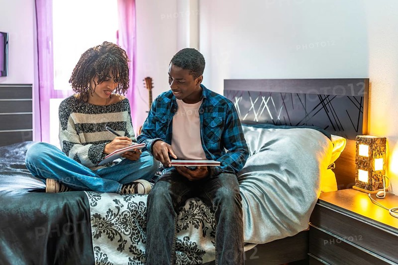 Two young students are sitting on a bed, collaborating on their studies, sharing ideas, and preparing for exams in a comfortable home environment