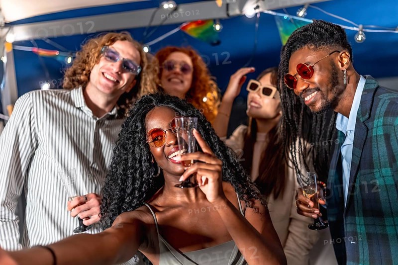 Beauty happy african woman holding champagne flute while taking selfie with elegant multi-ethnic friends in a terrace at night