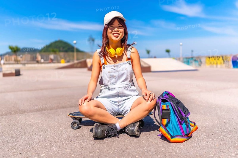 Asian woman sitting on skateboard smiling at camera in a public park