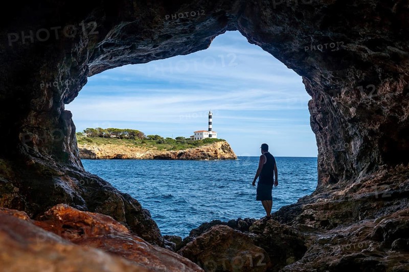 Tourist stands inside the arc natural cave in portocolom, mallorca, admiring the scenic view of a lighthouse across the blue waters