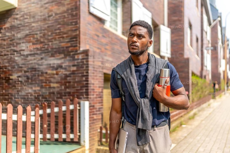 Young african entrepreneur walking the street with reusable coffee thermos and laptop