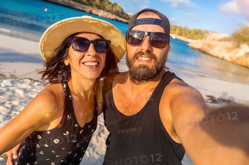 A man and woman are smiling for the camera on a beach. The man is wearing a black tank top and sunglasses, while the woman is wearing a straw hat