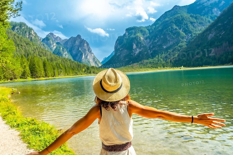 Woman wearing straw hat with open arms enjoying the pristine waters of lake dobbiaco and the majestic dolomites mountains in summer