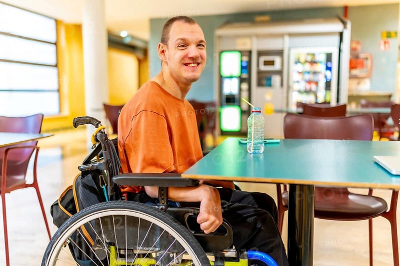 Three quarter length photo of a smiling caucasian man with disability sitting drinking water in the university cafeteria