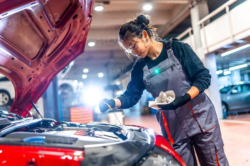 Young female mechanic wearing overalls and protective glasses is checking the oil level of a car engine in a garage
