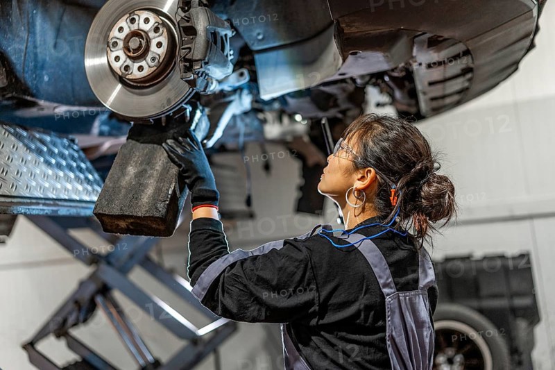 Skilled female mechanic working under a lifted car, inspecting and repairing the brake system in a professional workshop