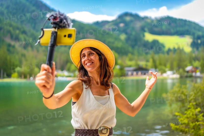 Smiling woman wearing straw hat vlogging with smartphone and external microphone at lake dobbiaco in the italian dolomites during summer holidays