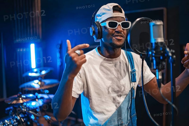 Talented african american artist wearing trendy sunglasses and headphones, passionately recording music in a modern studio, illuminated by blue lights