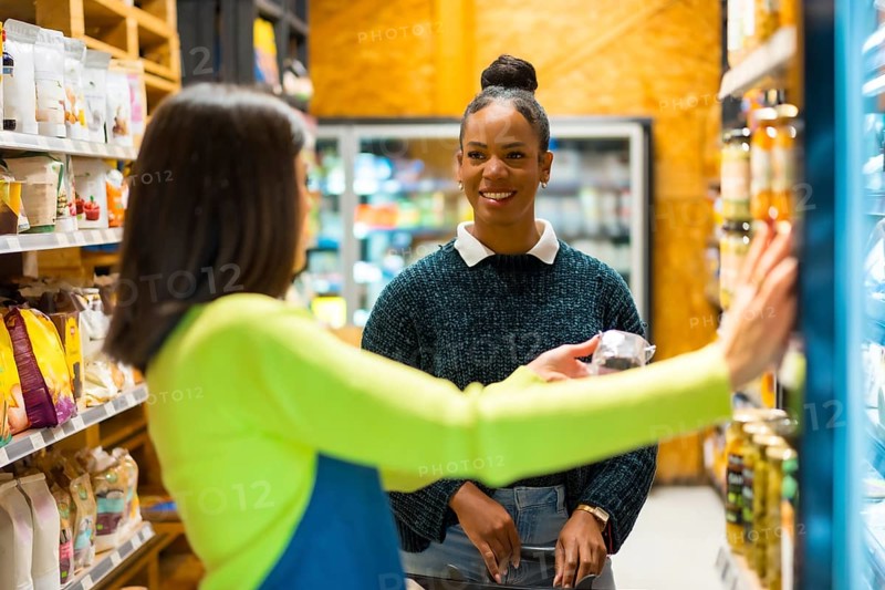 Smiling saleswoman advising customer choosing healthy food from refrigerated shelves, shopping in organic supermarket with natural and gluten free products