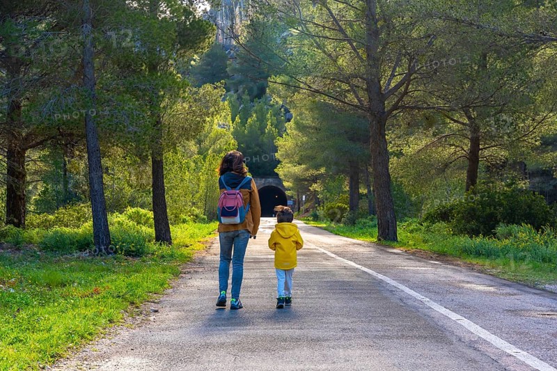 Mother and child enjoying a leisurely walk on a paved road amidst lush greenery and pine trees near a tunnel in la fontcalda sanctuary, gandesa, tarragona