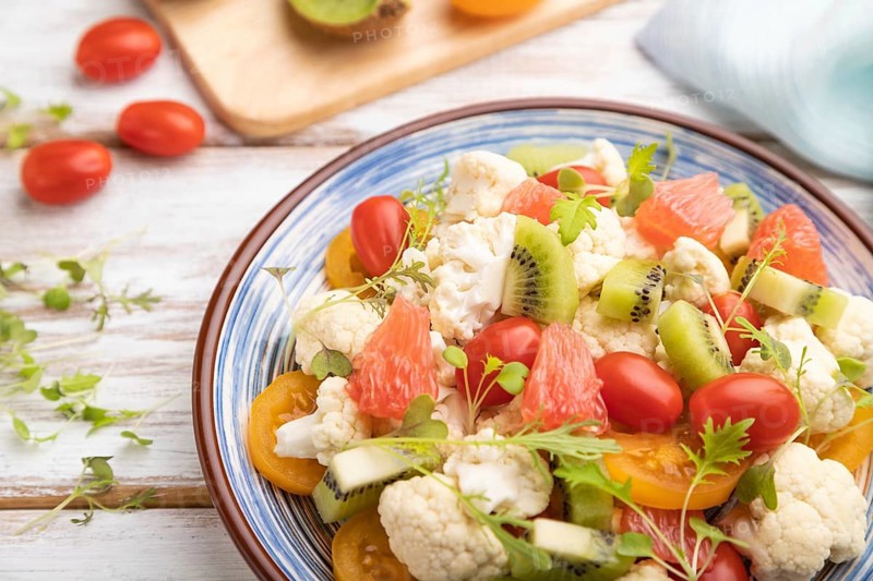 Vegetarian salad of cauliflower cabbage, kiwi, tomatoes, microgreen sprouts on white wooden background and blue linen textile. Side view, close up, selective focus