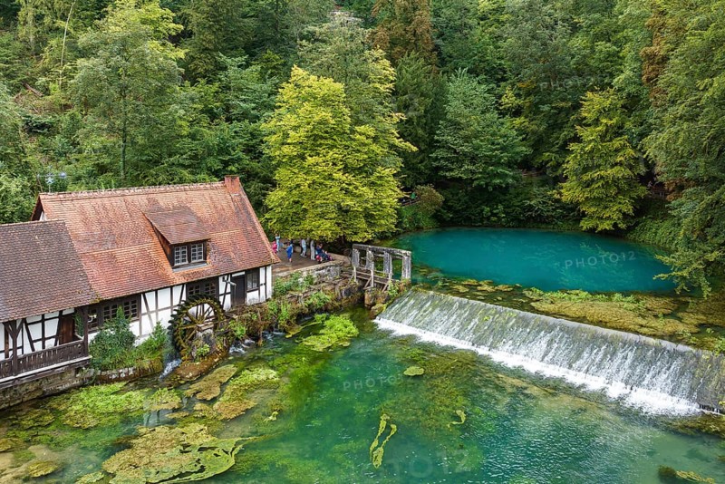 Blautopf Blaubeuren with industrial monument Hammerschmiede, source of the little river Blau in a landscape with forest. Karst spring, geotope and geopoint of the UNESCO Swabian Alb Geopark, tourist attraction. The popular excursion destination is now being thoroughly renovated and will therefore be closed to visitors until the end of 2028. Drone photo. Blaubeuren, Baden-Württemberg, Germany, Europe