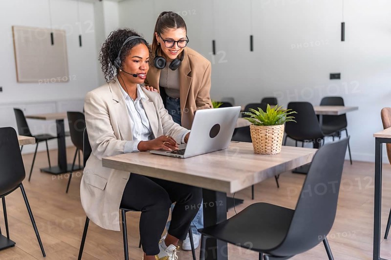 Two businesswomen collaborating on a laptop in a modern office, demonstrating teamwork and productivity in a professional setting