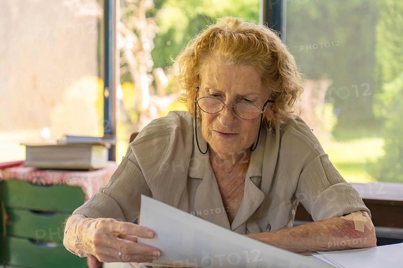 Focused elderly woman wearing glasses reading important documents while sitting at her home office desk, concentrating on paperwork and administrative tasks