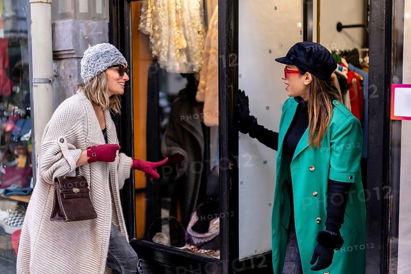 Young blonde girl enjoying shopping in the city and being attended by a shop assistant, looking at the window of clothing stores. Sunglasses and red gloves