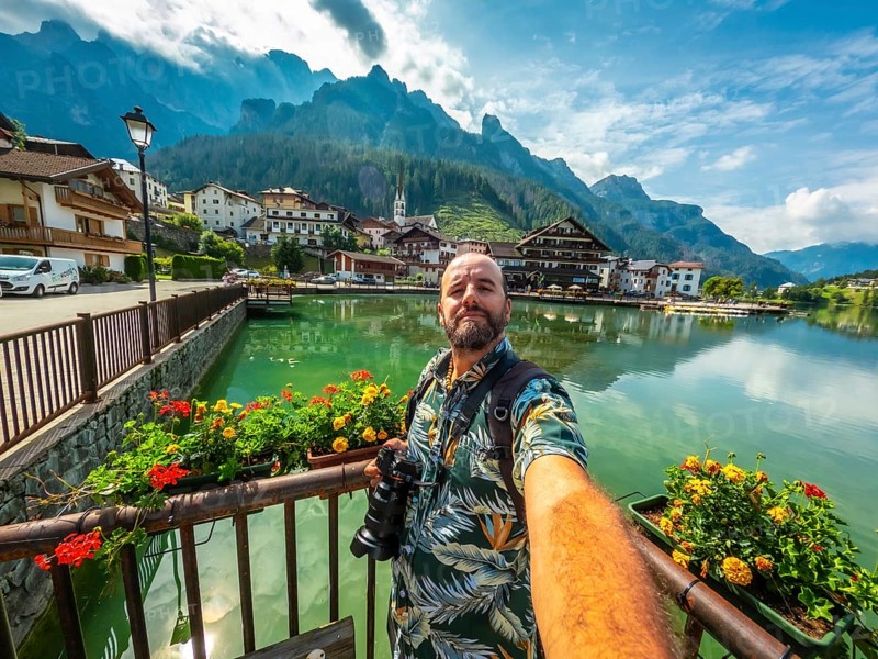 Photographer taking a selfie with alleghe lake and mountains in the background on a sunny summer day