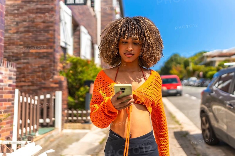 Frontal view of a beauty latin woman with wavy hair and colorful casual clothes using phone walking along a residential district